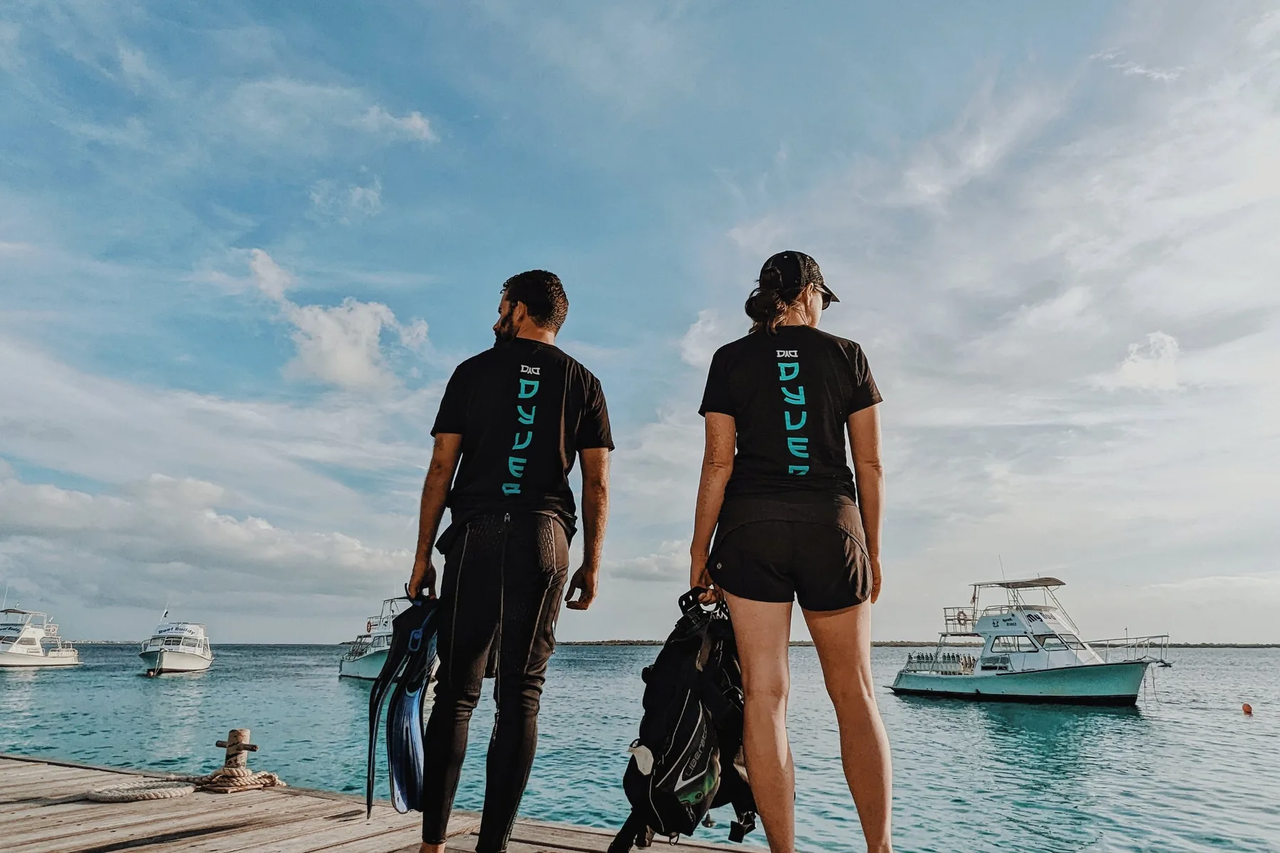 Two Dyver team members in matching Dyver shirts on a dock in Bonaire, holding fins and dive gear, looking out at the dive boats